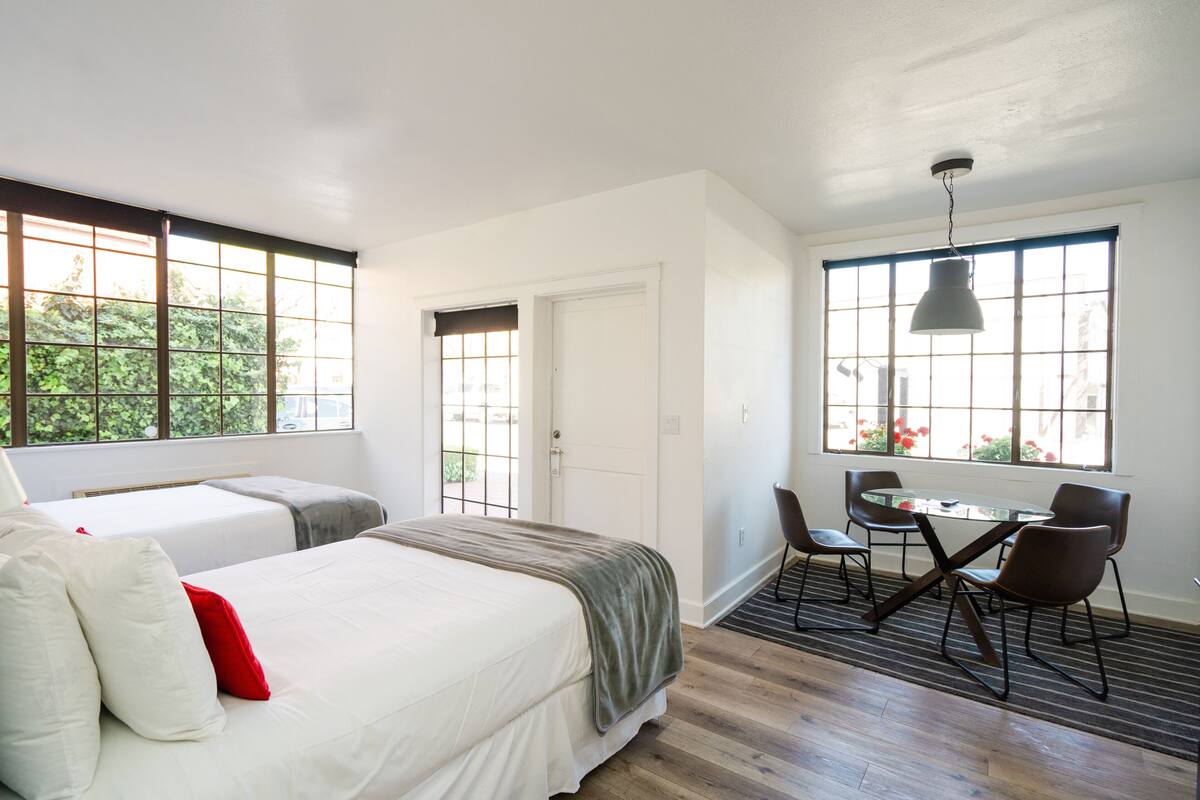 A hotel room with a bed, two bedside tables, a desk with chairs, and large windows with a view of greenery.