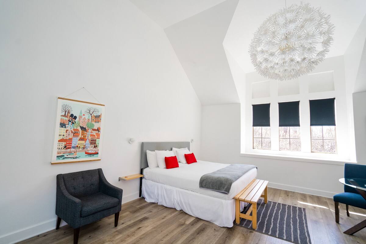 A bedroom with a bed, a chair, a small table, a window, and a wall-mounted artwork.
