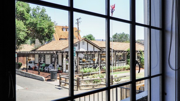 Une vue à travers une fenêtre ornée d’une grille métallique, dévoilant une scène de rue avec des bâtiments, un petit marché et un espace jardin.