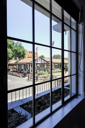 A view through a window with a metal grid, revealing a street scene with buildings, a small market, and a garden area. - Atterdag Inn (Solvang)