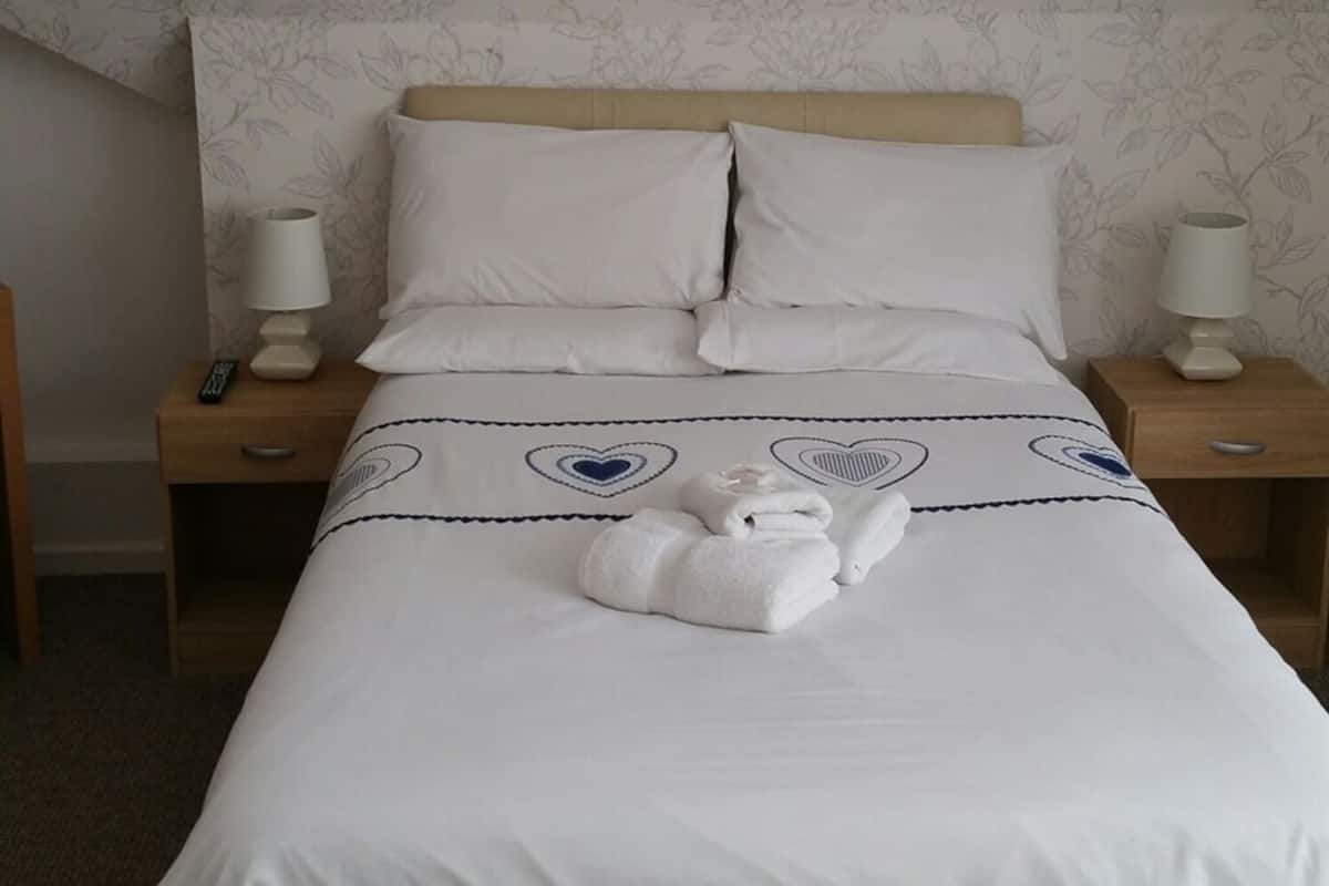 A neatly made bed with white linens, two pillows, and a folded towel. There are two bedside lamps on wooden nightstands.