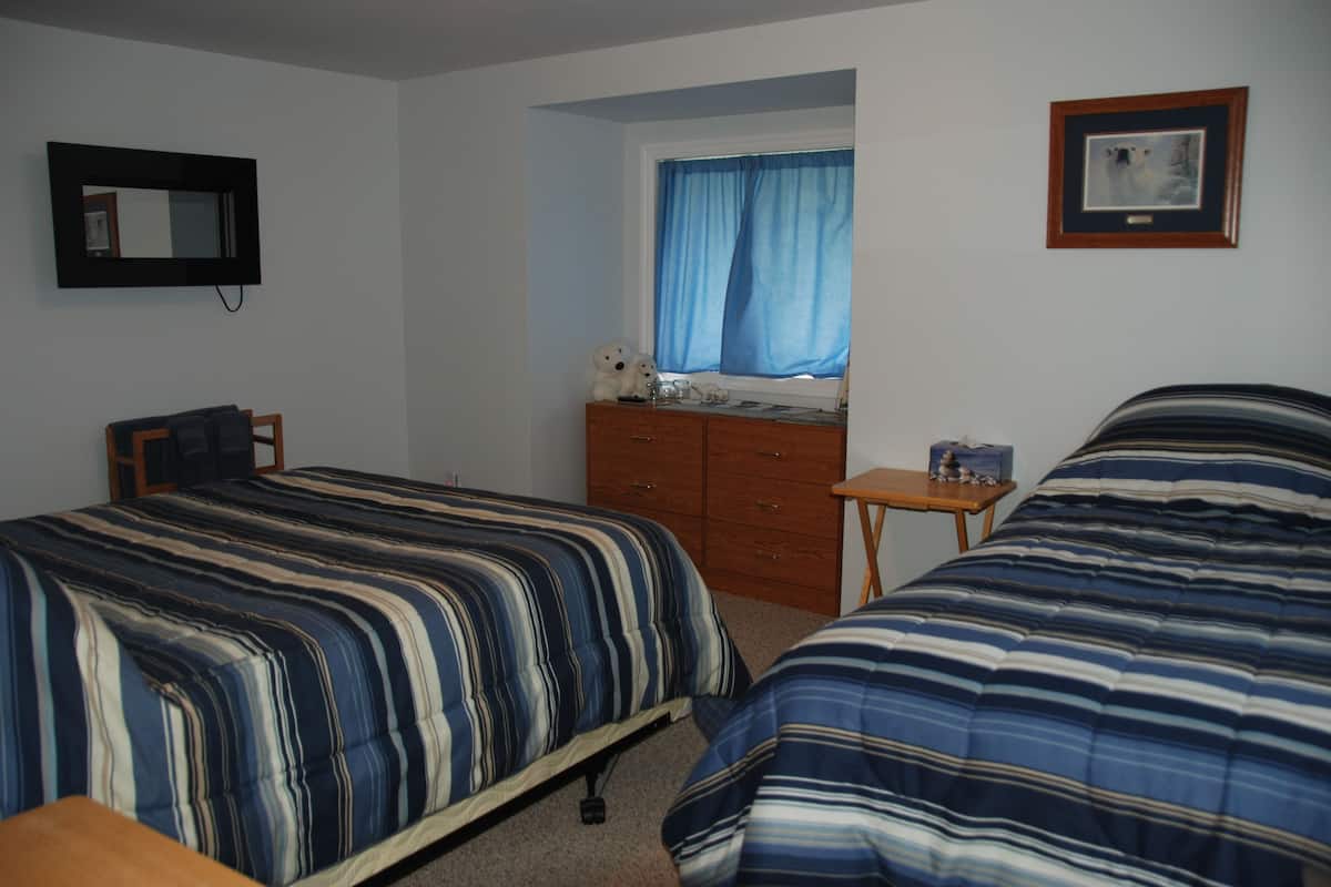 Un dormitorio con dos camas, un aparador, una silla, una mesita, una ventana con cortinas azules y un cuadro en la pared.