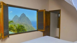 A bedroom with a bed, a window with a view of mountains and palm trees, and a door. - Piton Deck Villa (Soufrière)