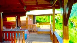 A wooden deck with a railing, a covered area, and a view of greenery. - Piton Deck Villa (Soufrière)