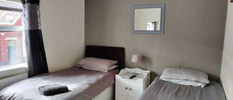 A hotel room with two single beds, a nightstand with a lamp, a mirror on the wall, and a window with curtains.