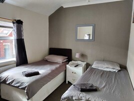 Habitación de hotel con dos camas individuales, una mesita de noche con lámpara, un espejo en la pared y una ventana con cortinas.