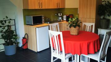 A compact kitchen with wooden cabinets, a microwave, and a coffee maker. There is a dining table with a red tablecloth and white chairs. A potted plant is on the table, and a fire extinguisher is near the kitchen.