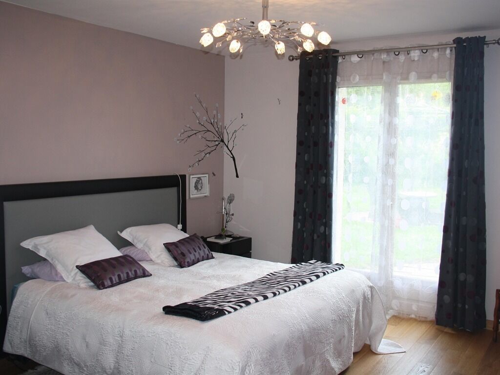A bedroom with a large bed, a nightstand, a wall-mounted lamp, and a window with curtains.