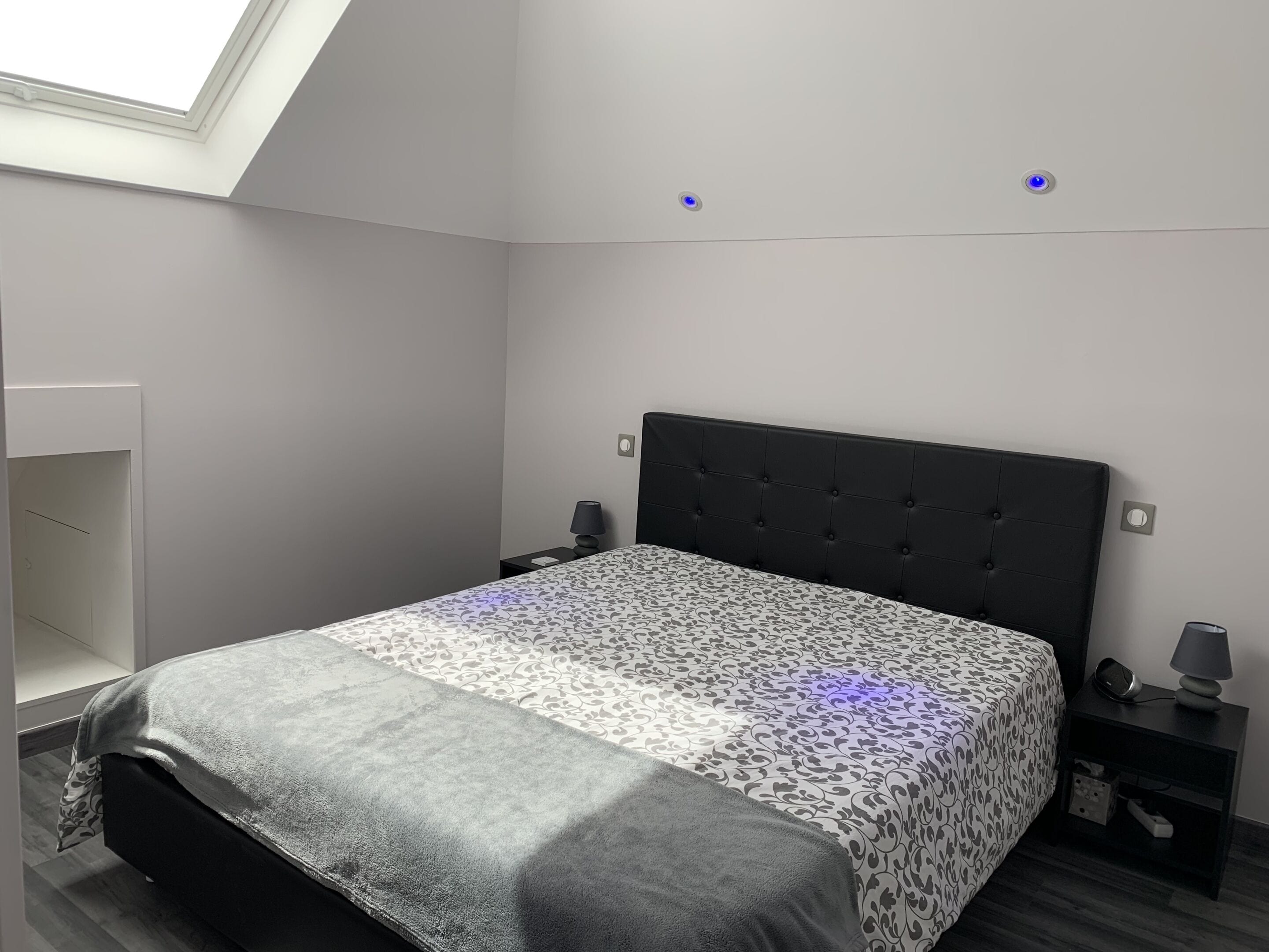 A bedroom with a bed, a nightstand, a lamp, and a skylight.