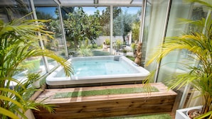 A hot tub with a wooden deck and artificial grass, surrounded by potted plants and a glass enclosure. - Villa Ambrosia (Narbonne)