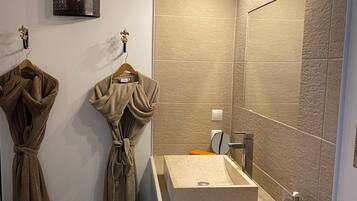 A bathroom with a beige tiled wall, a large mirror, a rectangular sink, and two robes hanging on hooks.
