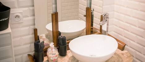 Un baño con pared de azulejos blancos, un espejo grande, un lavabo redondo y un mueble de tocador de madera.