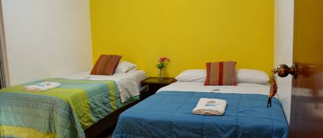 Una habitación con dos camas, cada una con una colcha de color diferente, una mesita con un florero y una puerta con pomo de latón.