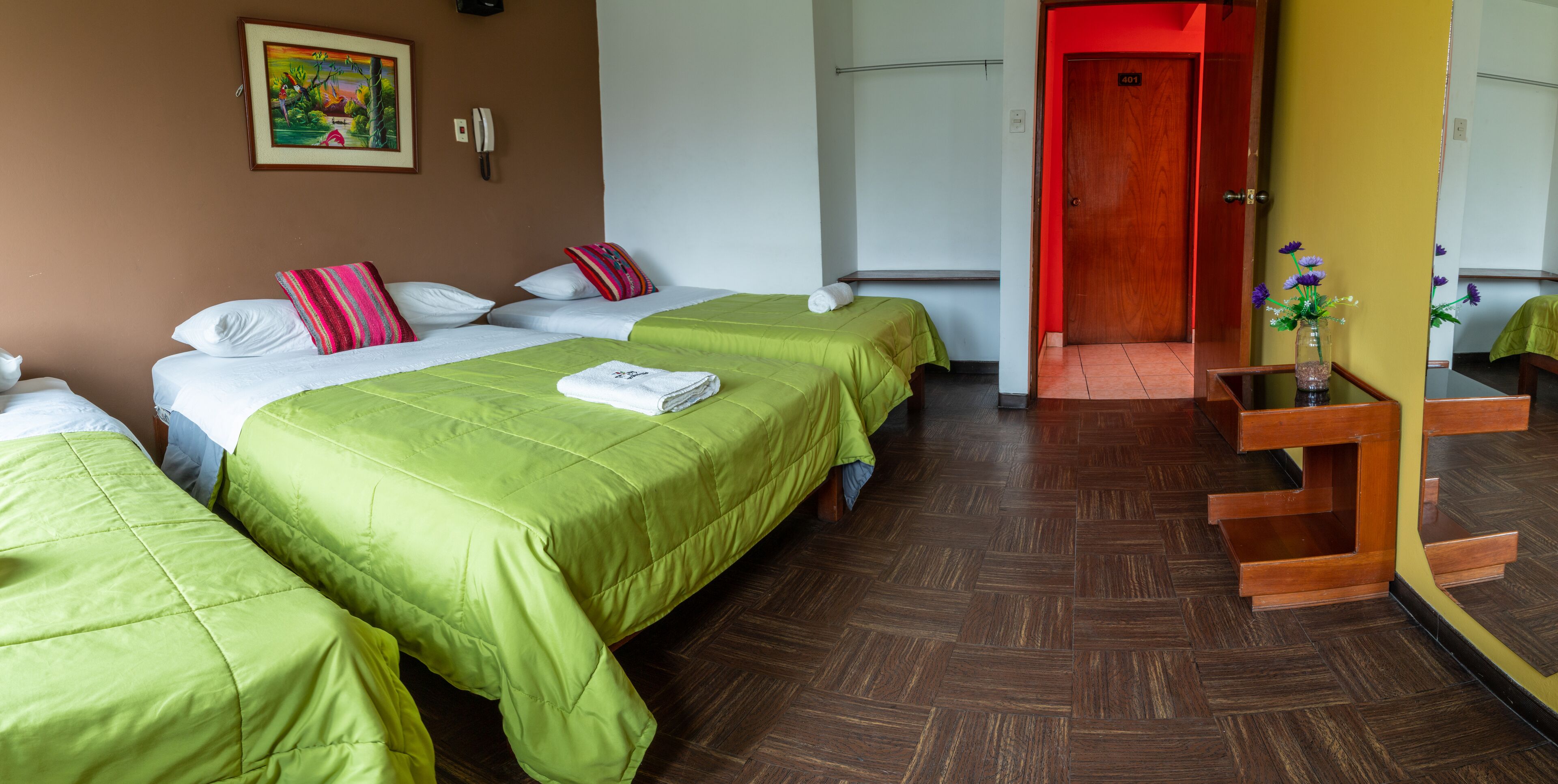 A hotel room with two beds, a wooden floor, a painting on the wall, and a wooden desk with a vase of flowers.