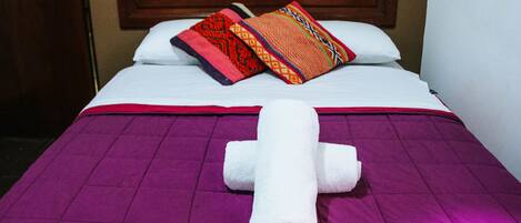 Una cama con una colcha morada, una manta blanca y una toalla blanca doblada en forma de 'X'. Hay dos almohadones decorativos sobre la cama.