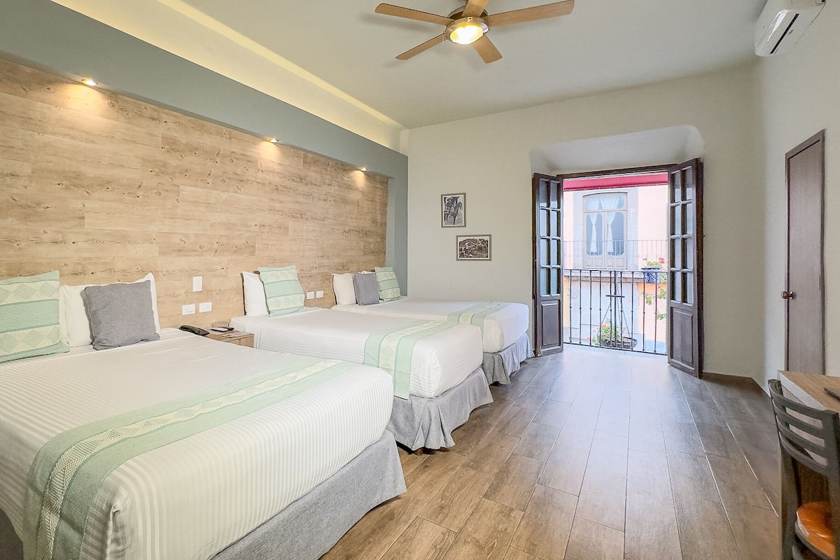 A hotel room with two beds, wooden flooring, a ceiling fan, and a balcony with a view.