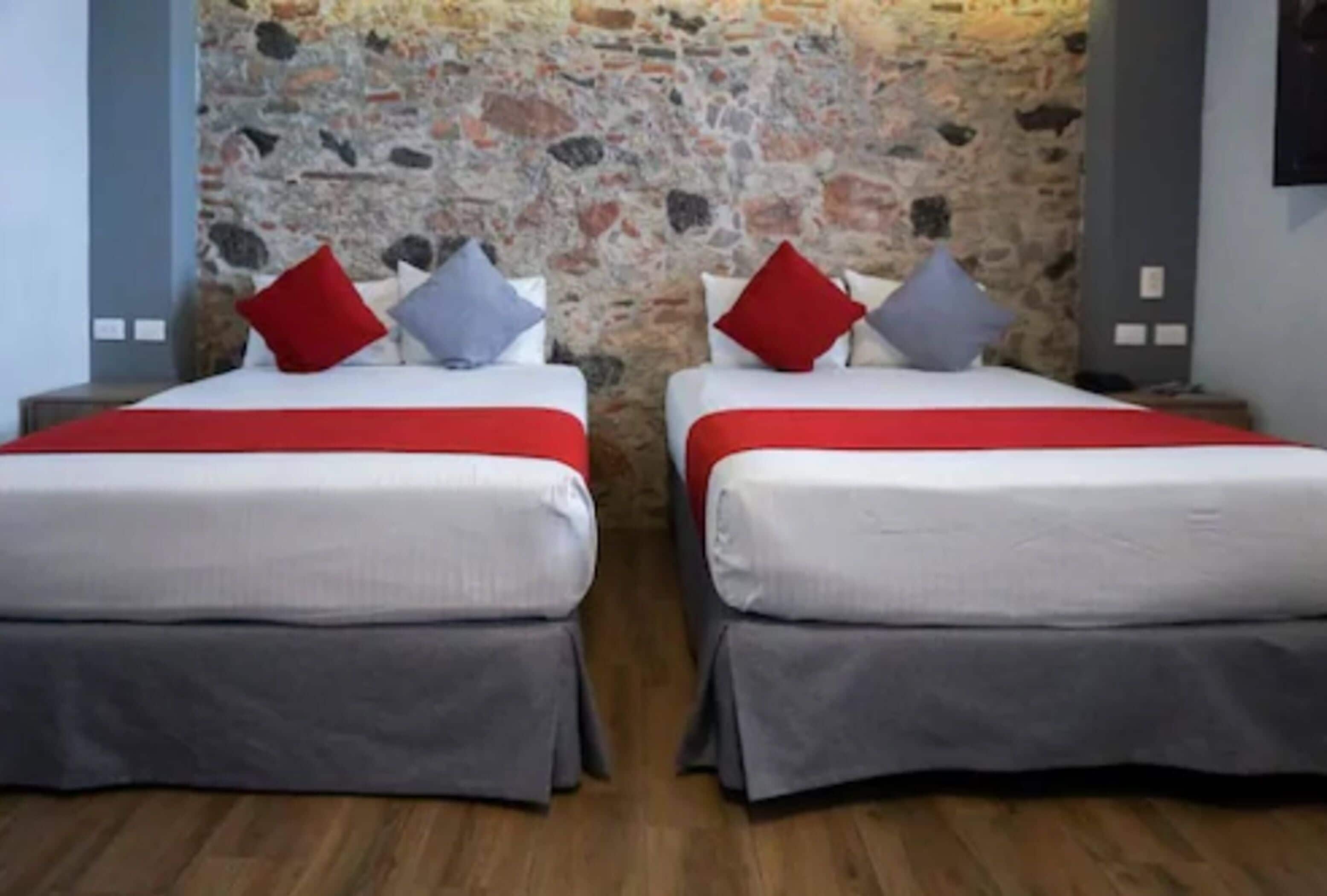 A double room with two beds, each with a red and grey bedspread, and a stone wall behind.