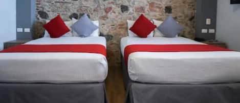 A double room with two beds, each with a red and grey bedspread, and a stone wall behind.