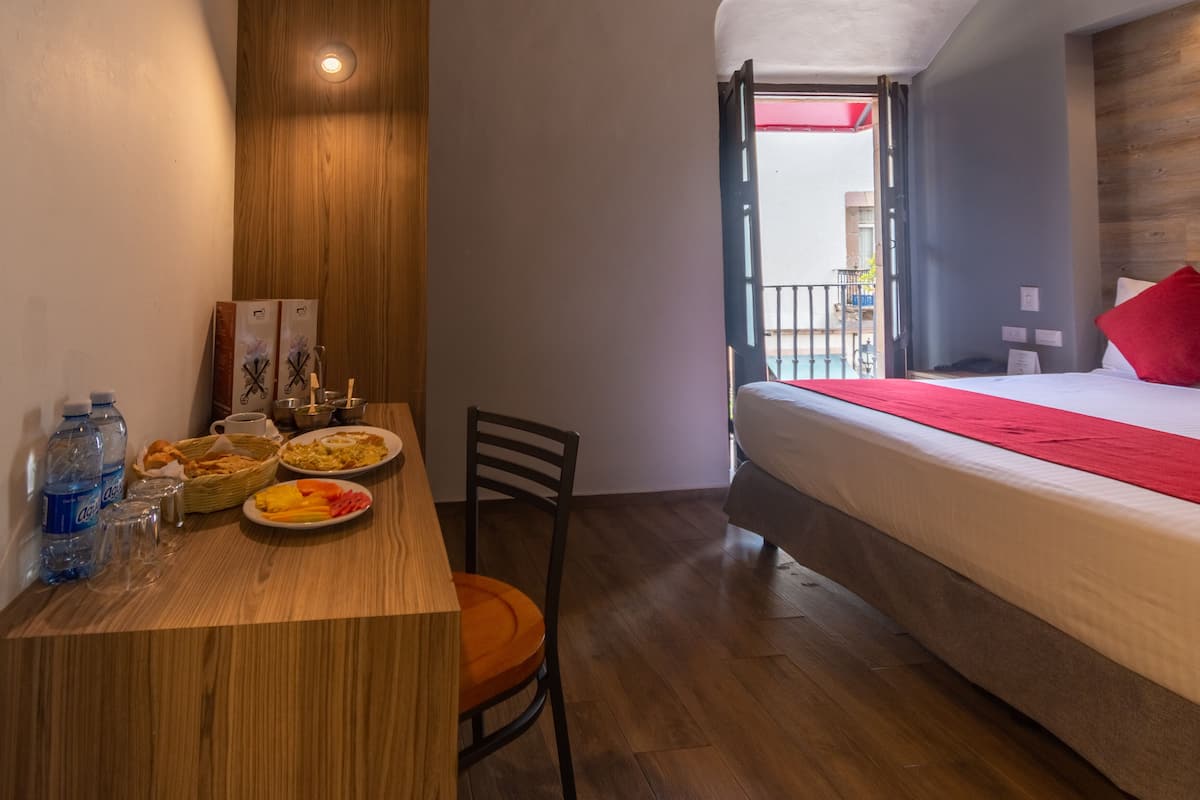 A compact hotel room with a wooden floor, a bed with a red pillow, a small table with breakfast items, and a chair.
