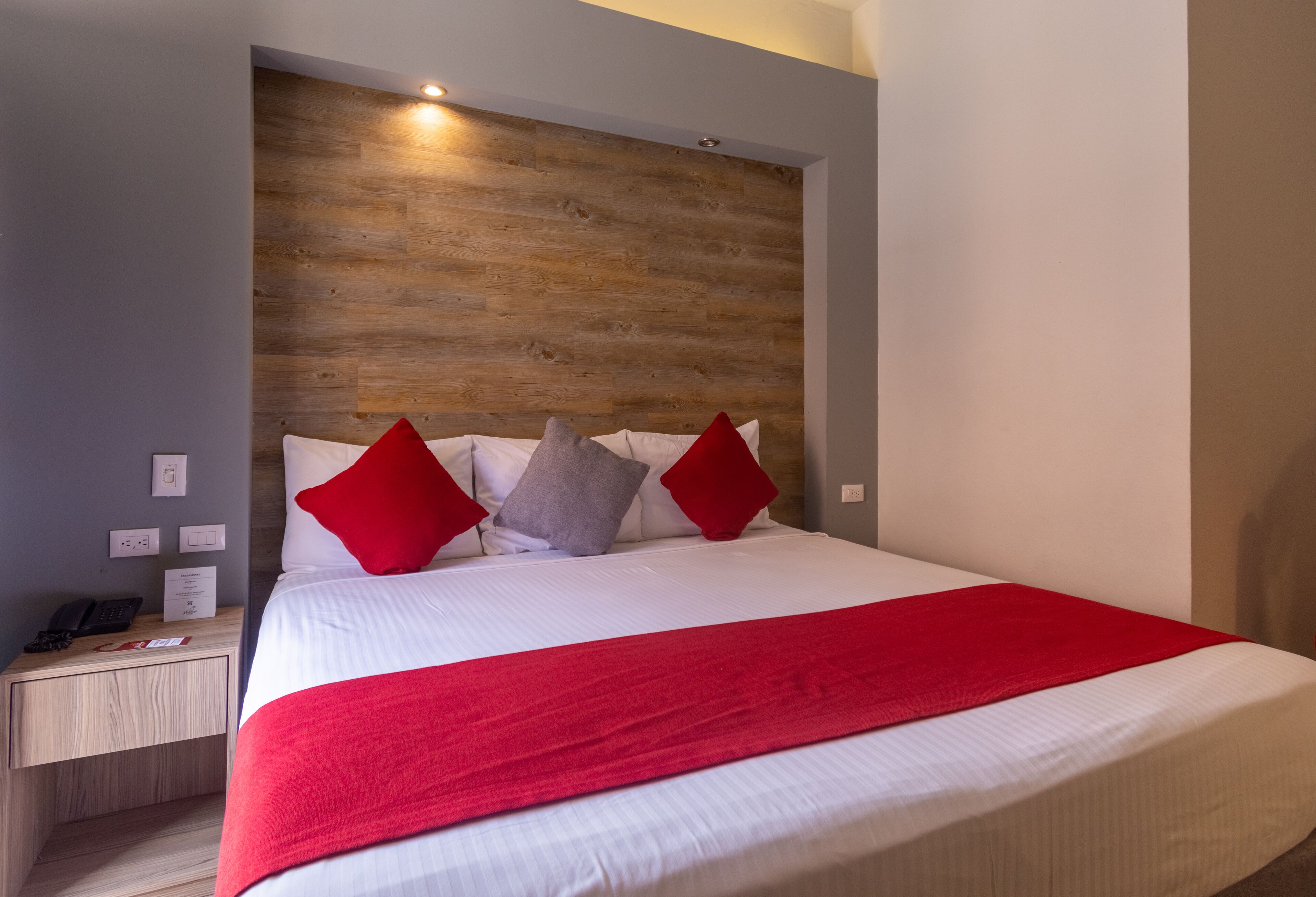 A neatly made bed with a red blanket and pillows, a wooden headboard, and a bedside table with a phone and a red object.