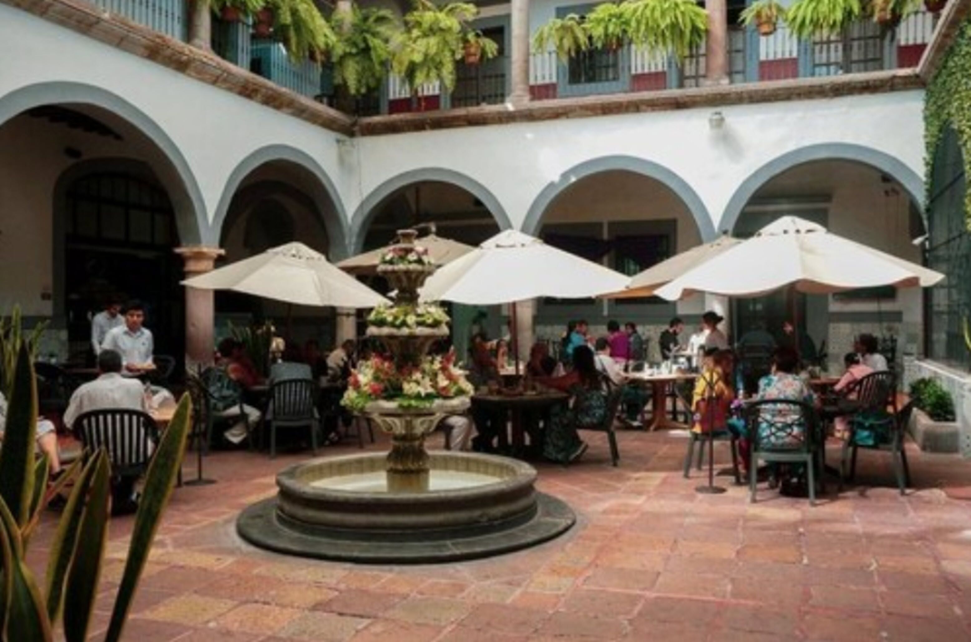 An outdoor courtyard with a central fountain, surrounded by tables and chairs under umbrellas.