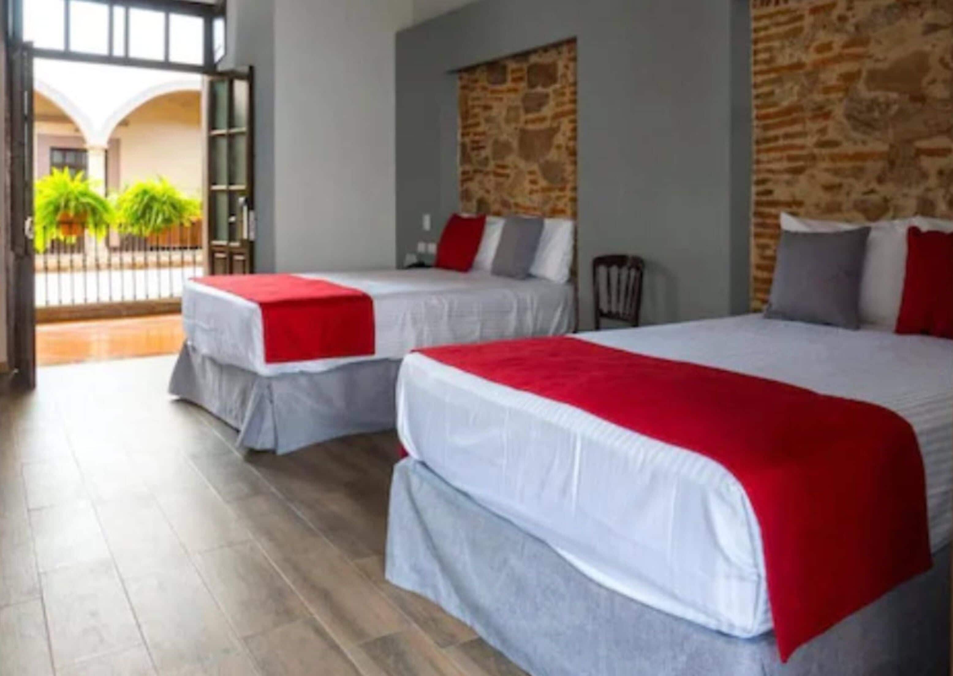 A hotel room with two beds, each with a red and white bedspread. The room has a stone wall and a view of a courtyard with plants.