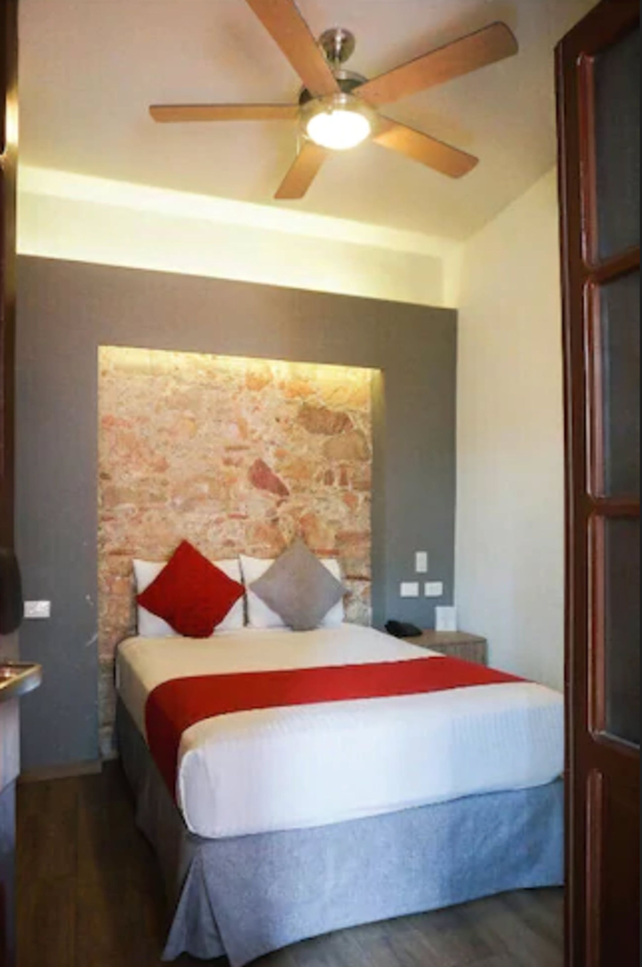Un dormitorio con cabecera de piedra, cama con colcha roja y blanca, ventilador de techo de madera y mesita de noche con un teléfono.