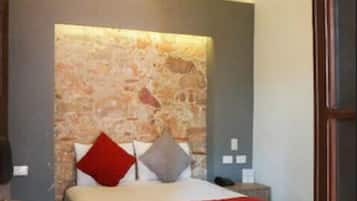 Un dormitorio con cabecera de piedra, cama con colcha roja y blanca, ventilador de techo de madera y mesita de noche con un teléfono.