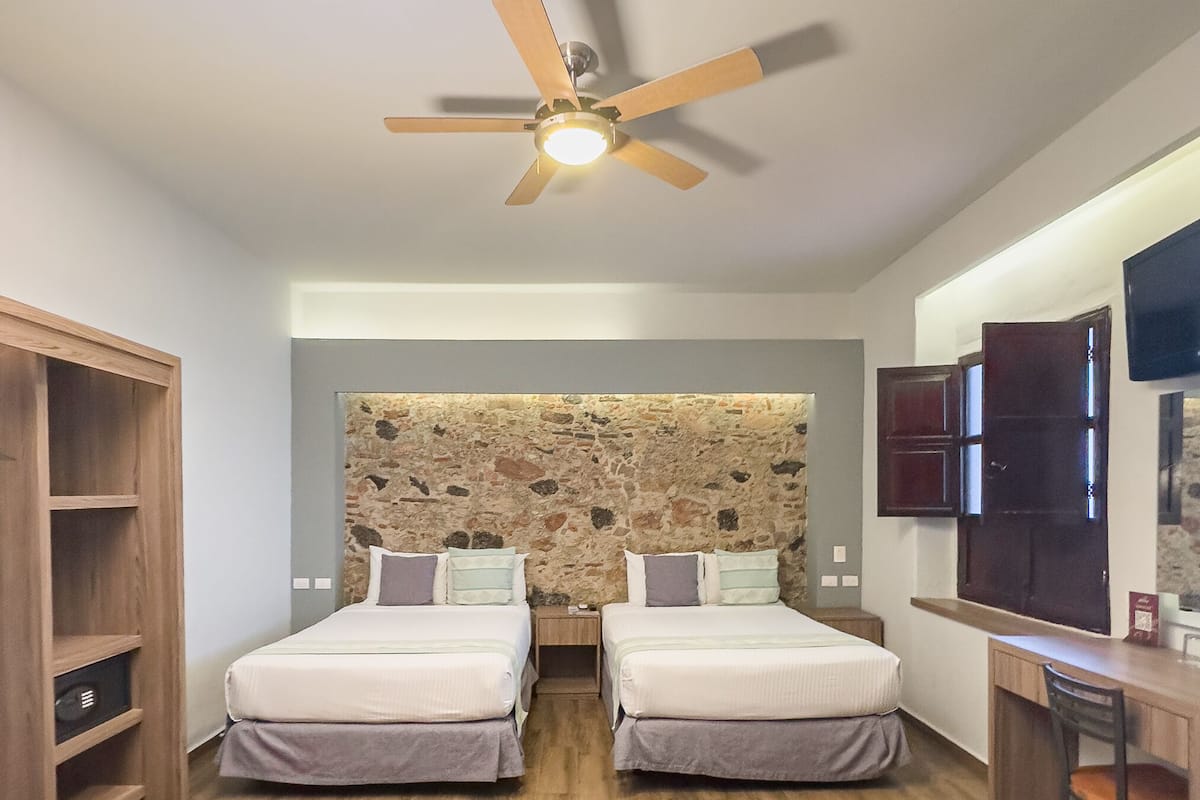 A hotel room with two beds, a wooden wardrobe, a desk, a television, and a ceiling fan.