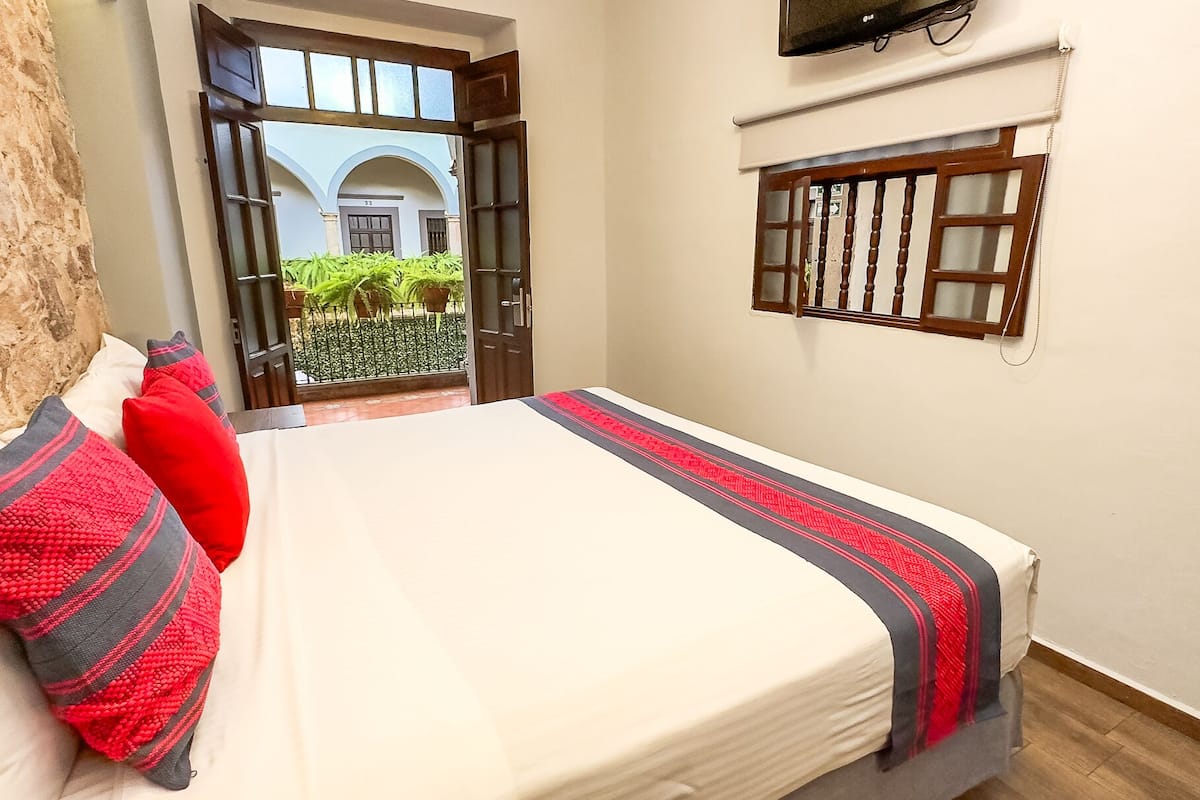 A bedroom with a bed, pillows, a window, and an open door leading to a courtyard.