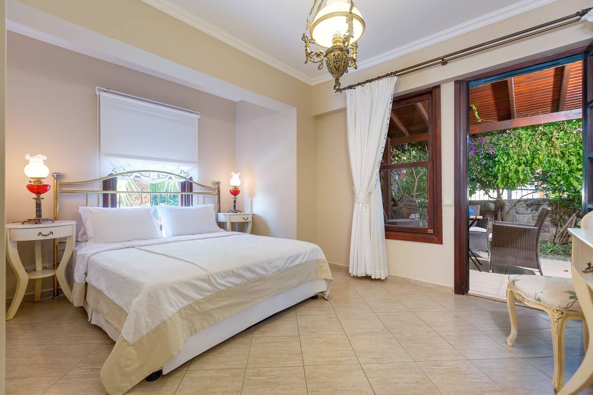A bedroom with a bed, two bedside tables with lamps, a window with curtains, and a view of an outdoor patio.