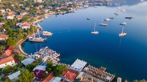 Vista aérea de un pueblo costero con un puerto deportivo lleno de barcos, una zona de muelles con tumbonas y edificios residenciales con tejados rojos.