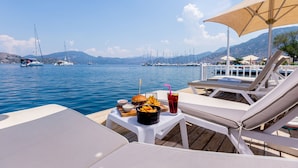 Una terraza con sillones, una mesa con comida y bebidas, y vista al mar con barcos y montañas al fondo.