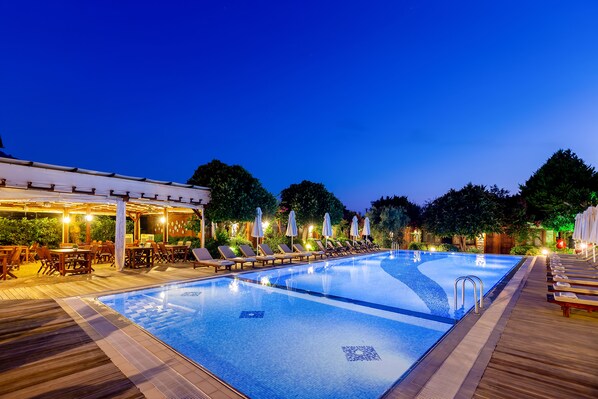 Un área de piscina con sillones de descanso y sombrillas, rodeada de terrazas de madera y árboles.