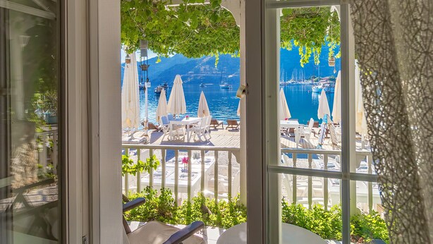 Vista desde una ventana que muestra una terraza con mesas y sillas, barandilla y un cuerpo de agua con barcos.