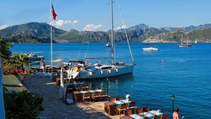 Un espacio de restauración frente al mar, con mesas y sillas, un velero anclado cerca y montañas al fondo.