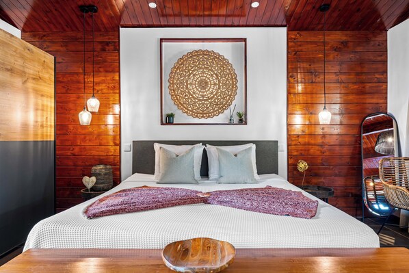 A modern bedroom with a wooden ceiling, a large bed with white bedding, two bedside lamps, and a decorative wall piece.