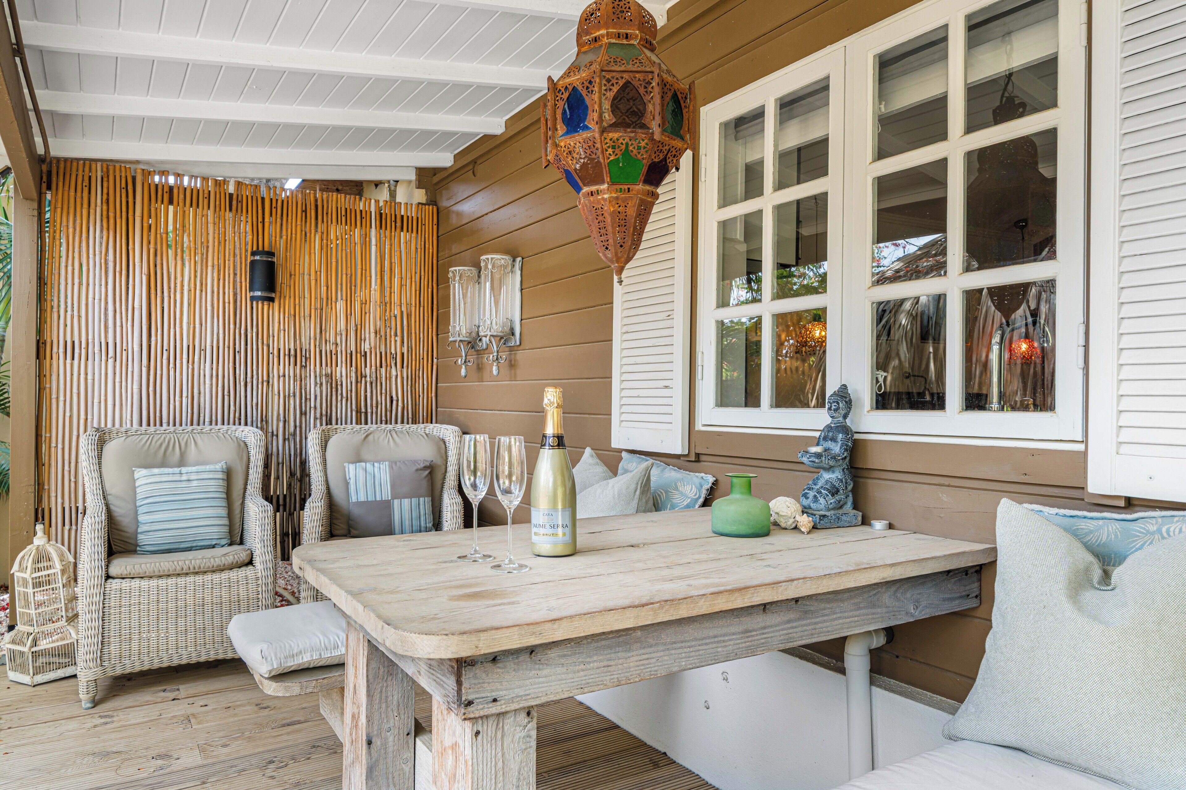 A wooden deck with wicker furniture, a table with a bottle and glasses, and a decorative hanging light.