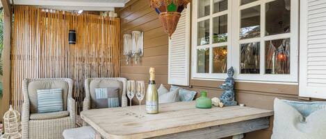 A wooden deck with wicker furniture, a table with a bottle and glasses, and a decorative hanging light.