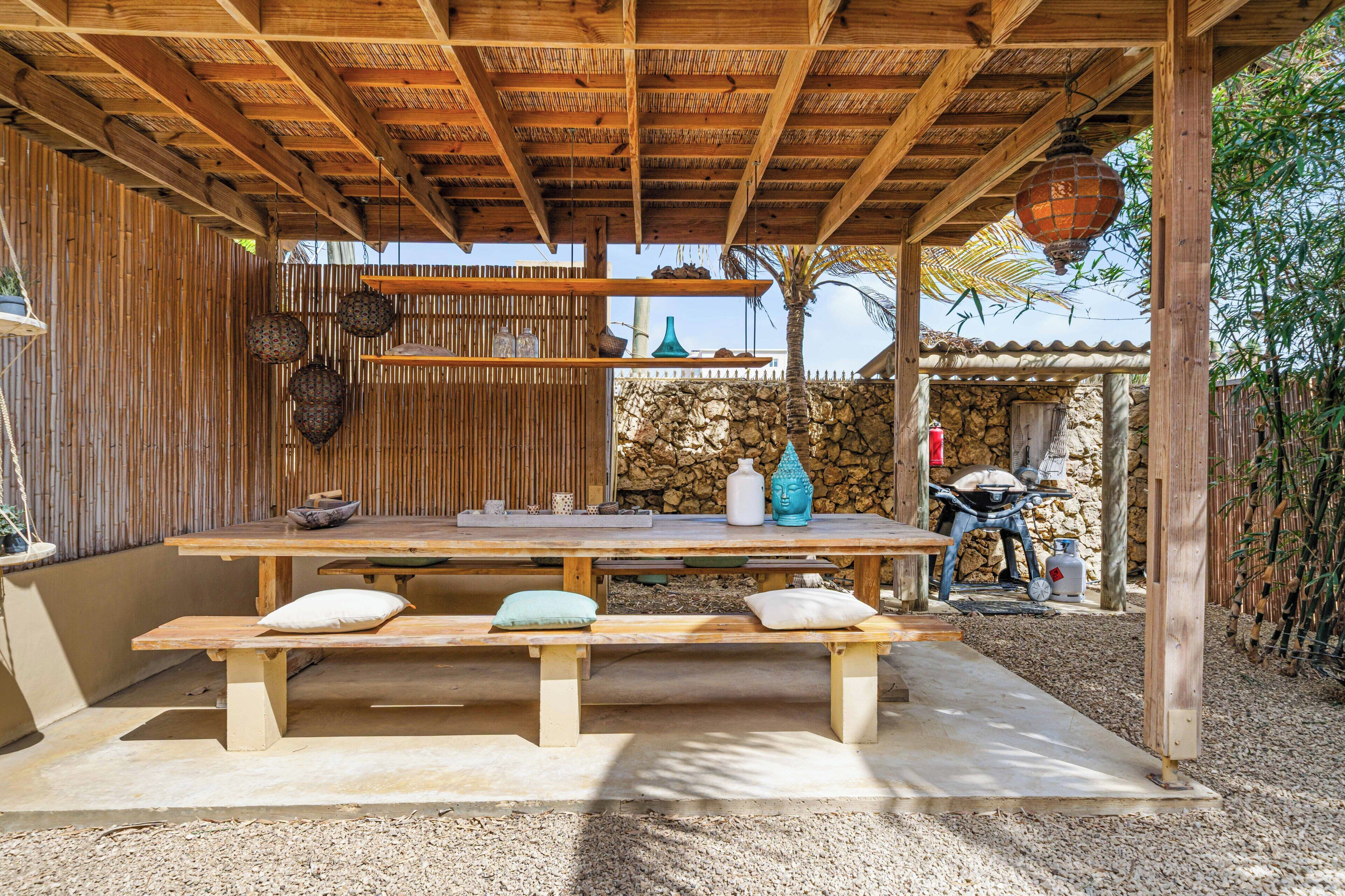 An outdoor wooden pergola with a table and benches, surrounded by a bamboo fence and a stone wall.