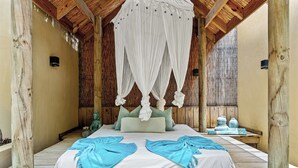 Quarto com cama, pilares de madeira, dossel e uma pequena mesa com objetos decorativos.