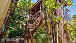 Uma escadaria de madeira leva a um edifício com fachada amarela, cercado por vegetação tropical exuberante.