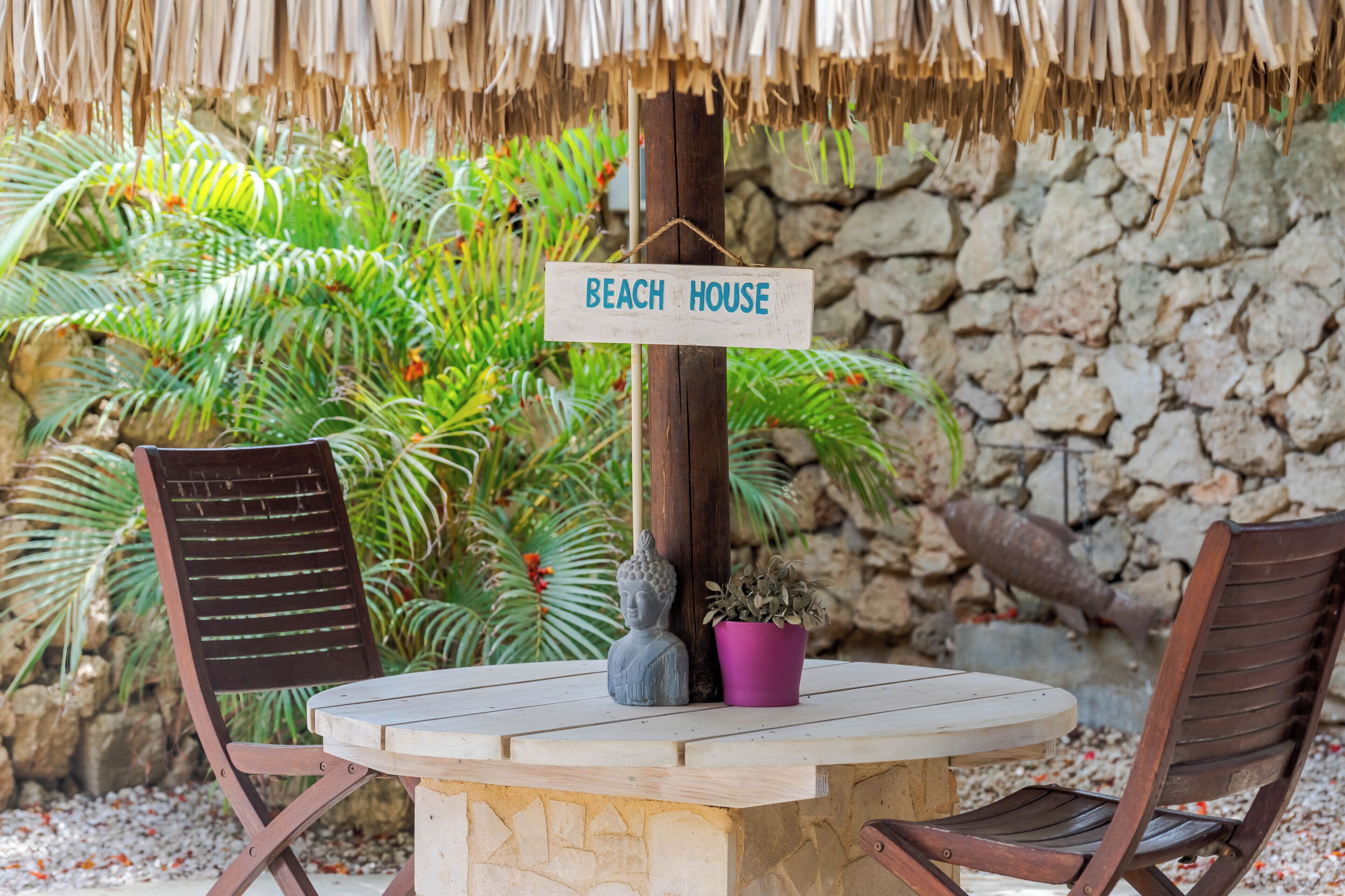 A round table with two wooden chairs, a sign reading 'BEACH HOUSE,' a Buddha statue, and a potted plant.