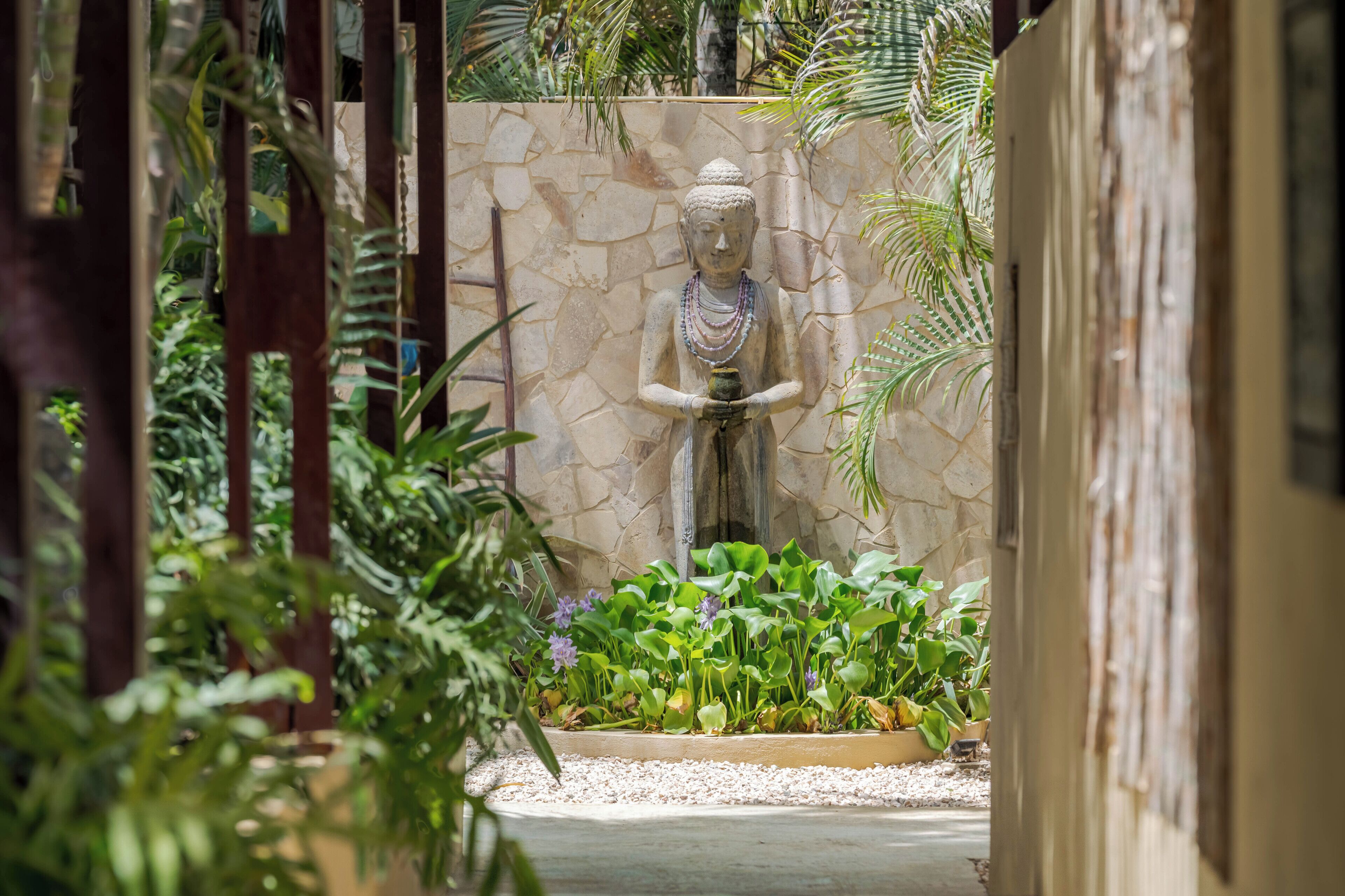 A statue in a garden with a stone wall, surrounded by various plants and a pathway.