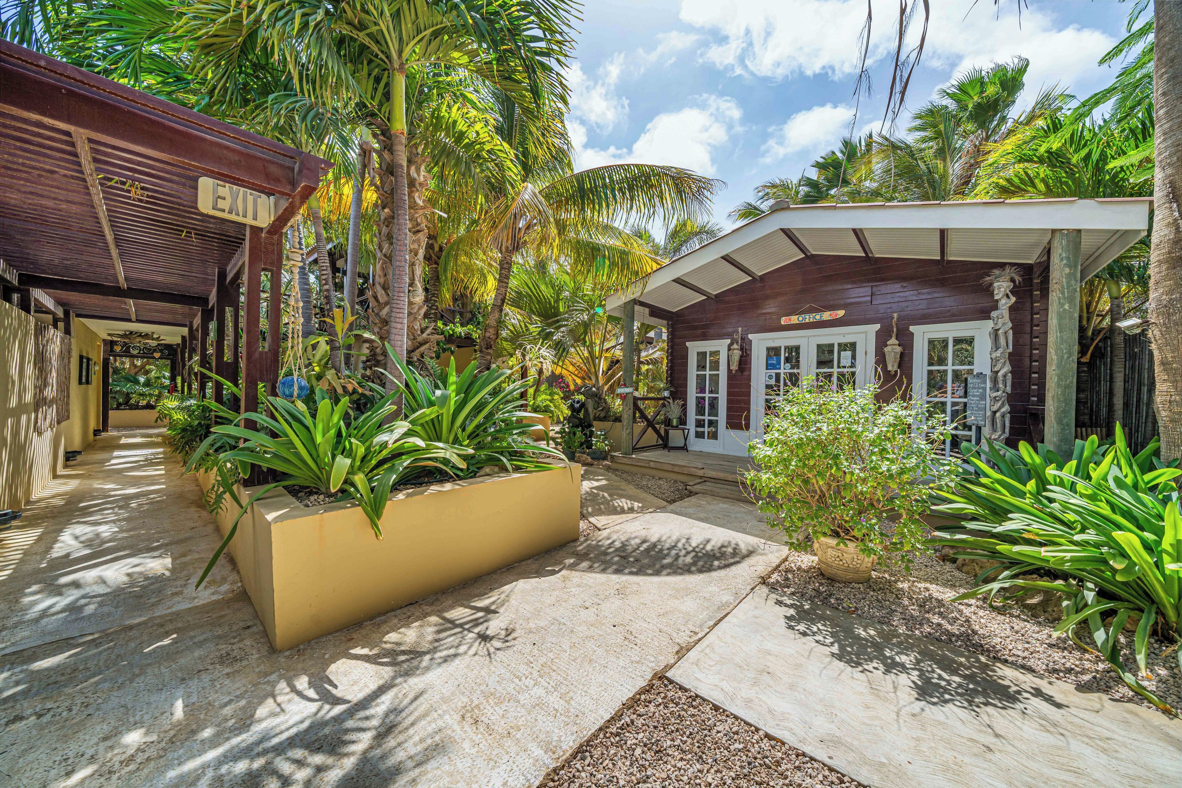 A covered outdoor area with a sign reading 'EXIT,' surrounded by tropical plants and a building with a white facade.