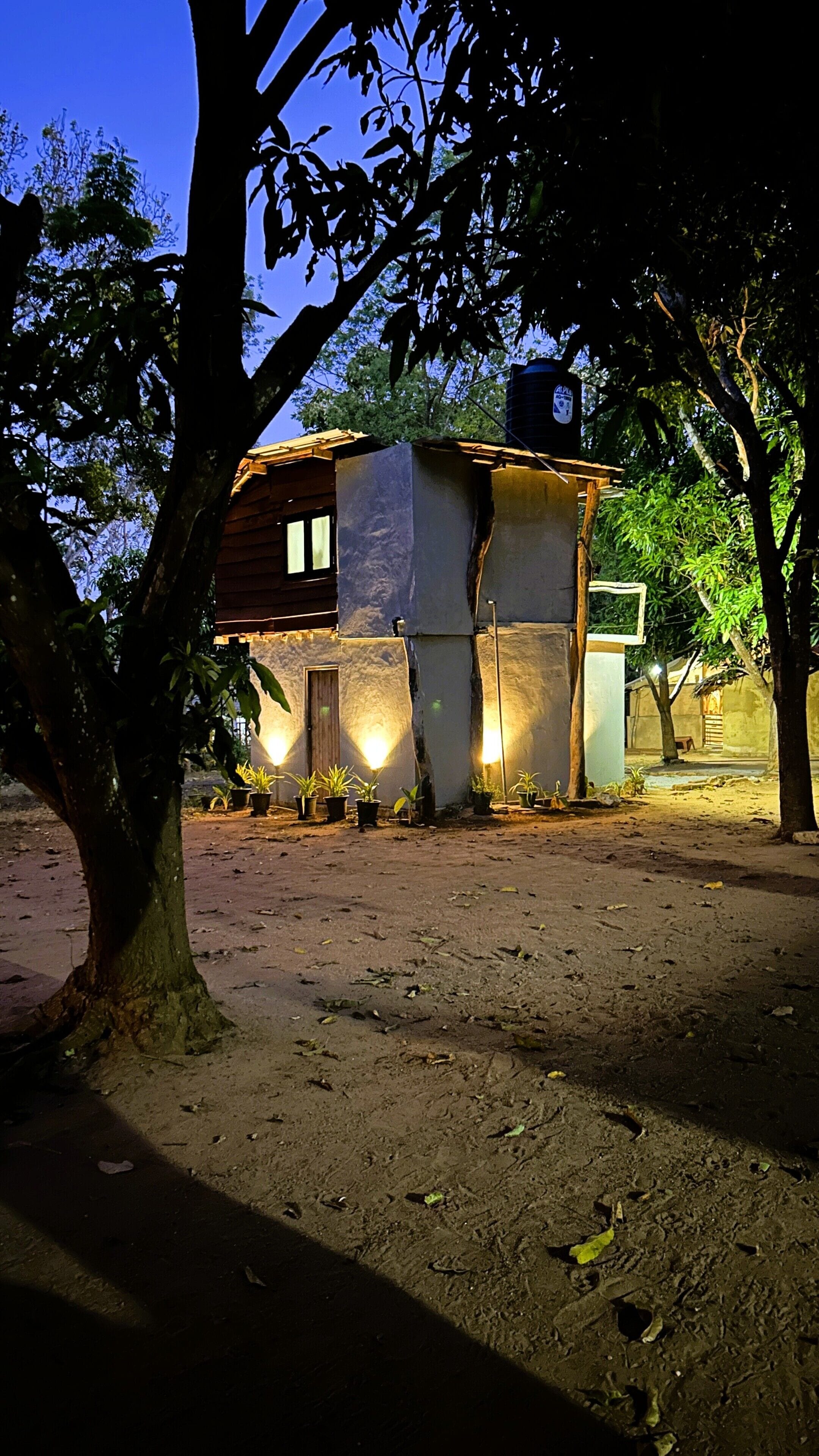 A house with a dirt ground, surrounded by trees, and has a water tank on the roof.