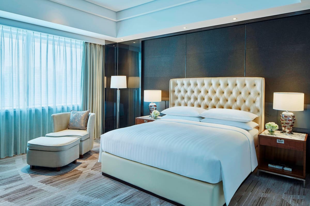 Una habitación de hotel moderna con una cama grande, una zona de estar con una silla y reposapiés, una mesita de noche con lámparas y vistas de la ciudad a través de grandes ventanales.