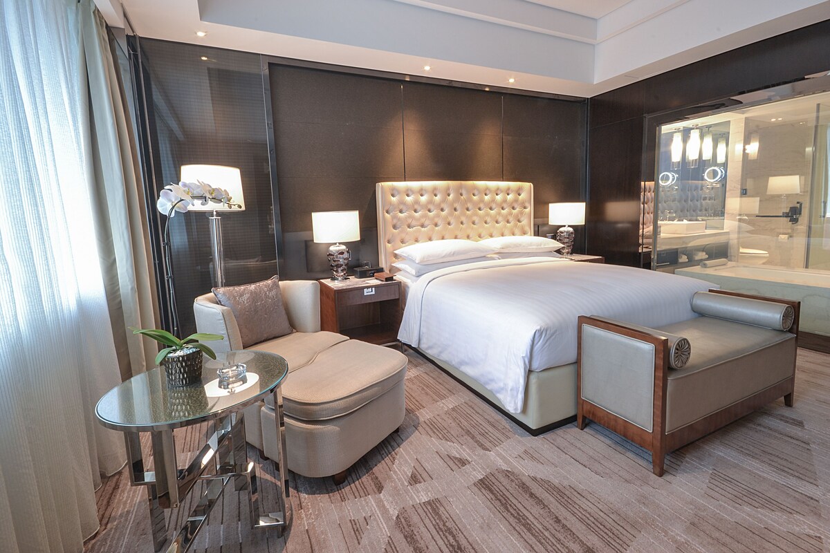 Une chambre d’hôtel moderne avec un grand lit, un coin salon doté d’une table en verre, une petite table avec une plante et une grande fenêtre avec des rideaux.