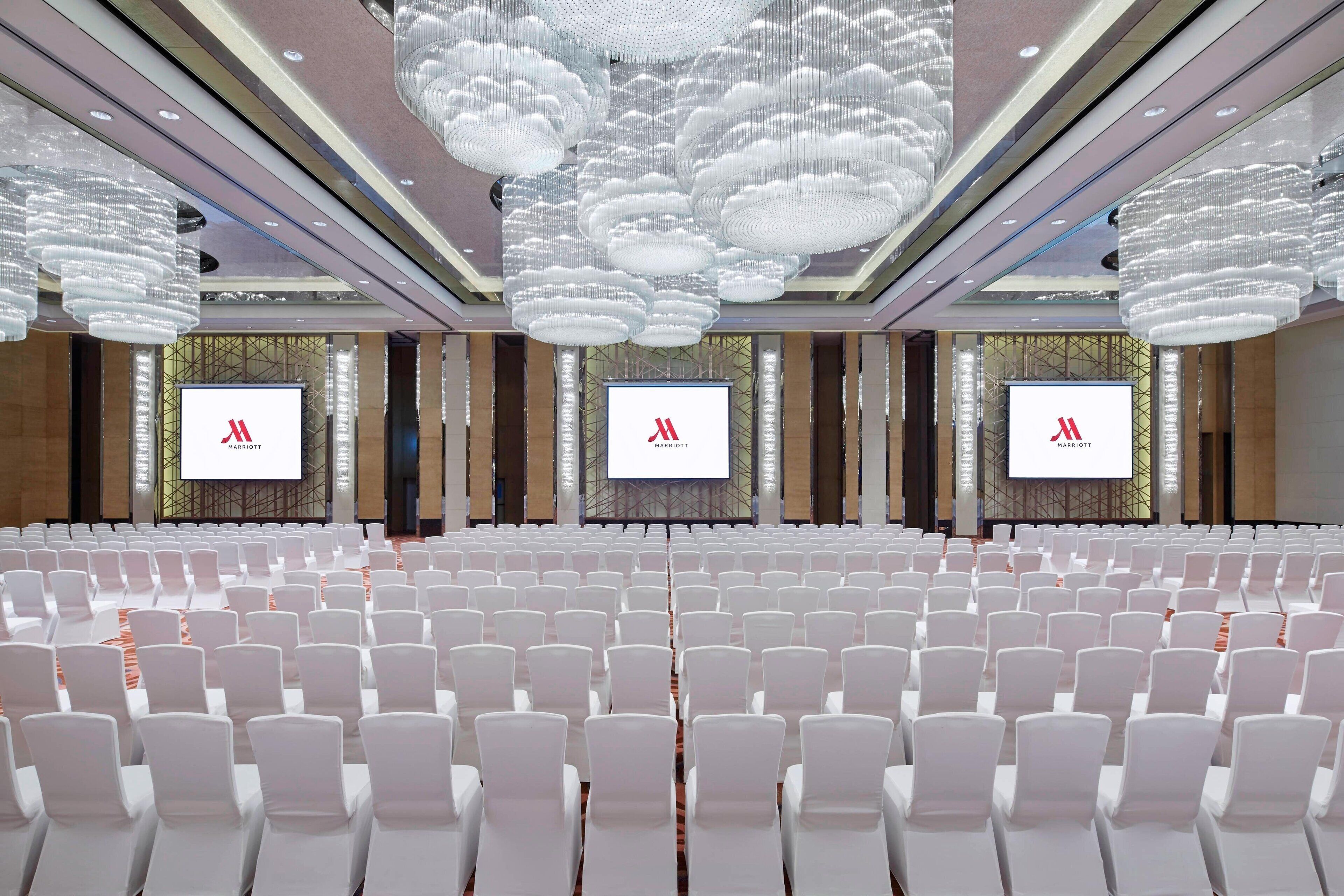 Une salle de conférence avec des rangées de chaises blanches, de grands lustres et des écrans affichant le logo de Marriott.