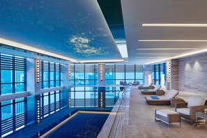 Un espace piscine intérieur moderne, doté de grandes fenêtres, de chaises longues et d’une piscine centrale.